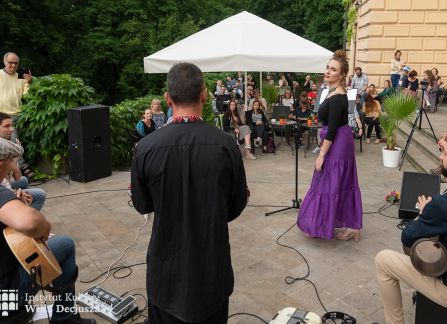 fotografia. dni wyszehradzkie w willi decjusza. koncert zespołu balkan artz. kobieta w długiej fioletowej spódnicy śpiewa. za nią na kamiennym tarasie willi w półkolu siedzą pozostali członkowie zespołu z instrumentami.